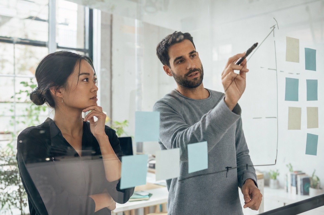 Two marketing strategists planning at a glass whiteboard covered in sticky notes and an upward arrow diagram